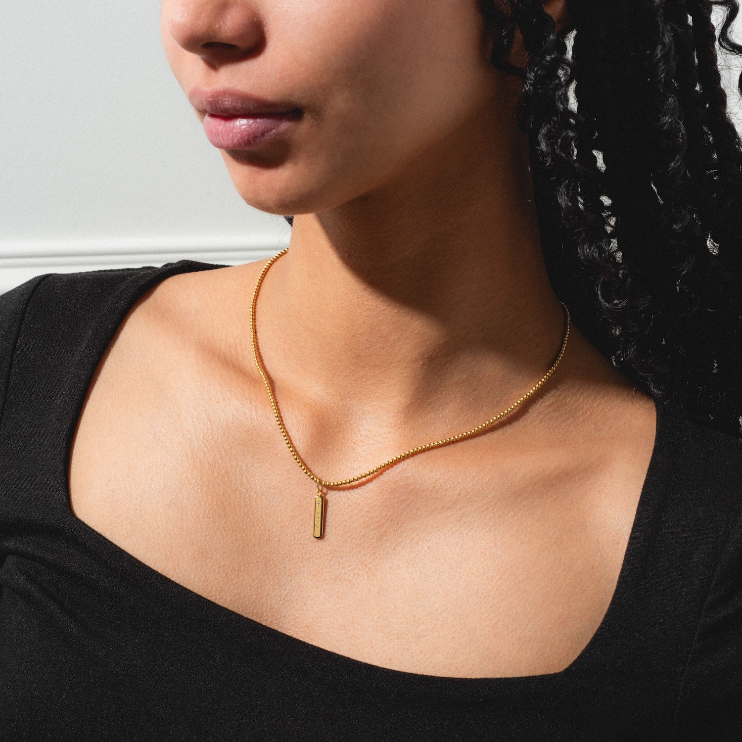 A woman with curly hair wears a black top and a simple gold necklace featuring the Tiny Fuck Charm with a vertical bar pendant, shown from the shoulders up against a light background.