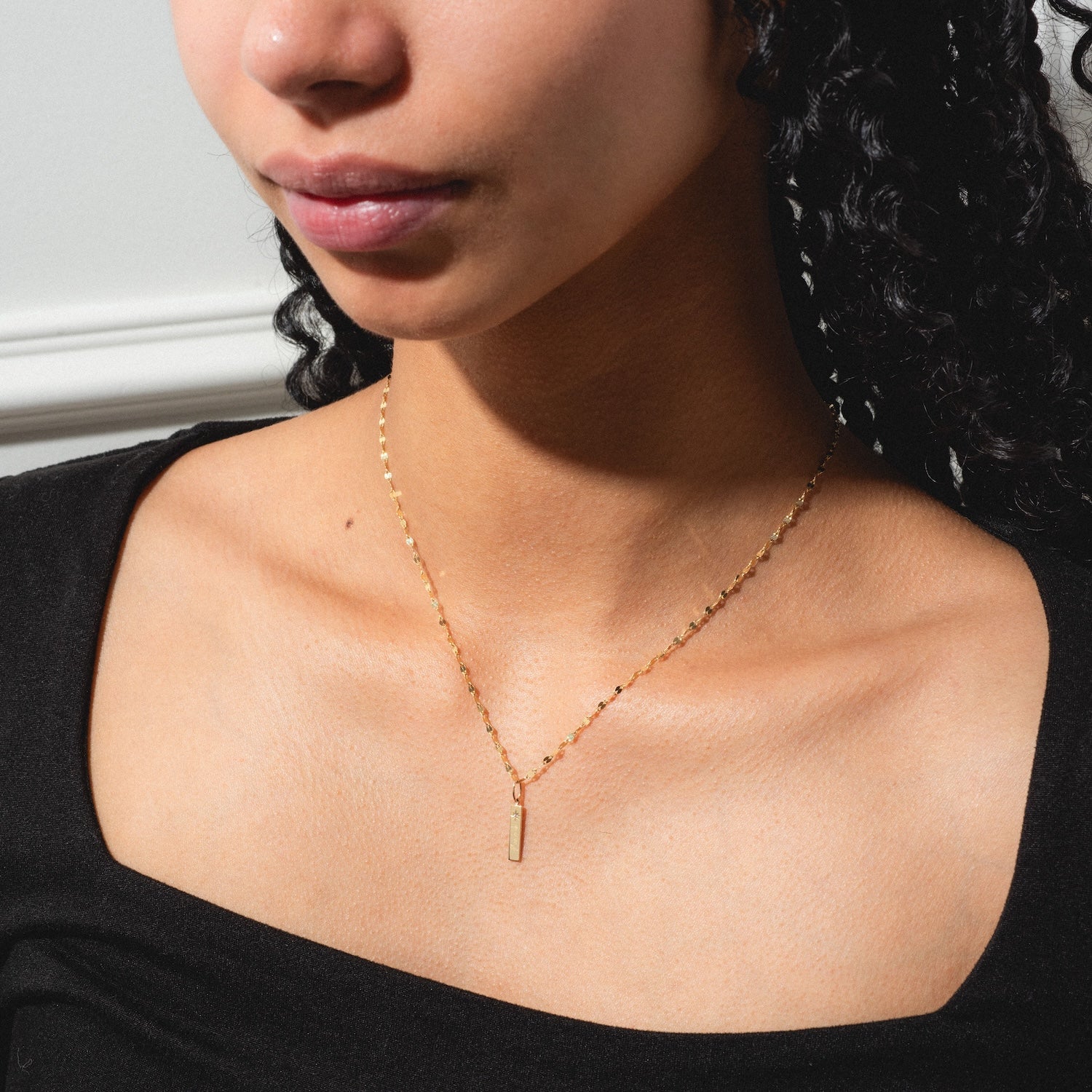 A woman wearing a black top and the Solid Gold Tiny Fuck Charm necklace with a vertical bar pendant, shown from the shoulders up with curly hair partly visible.