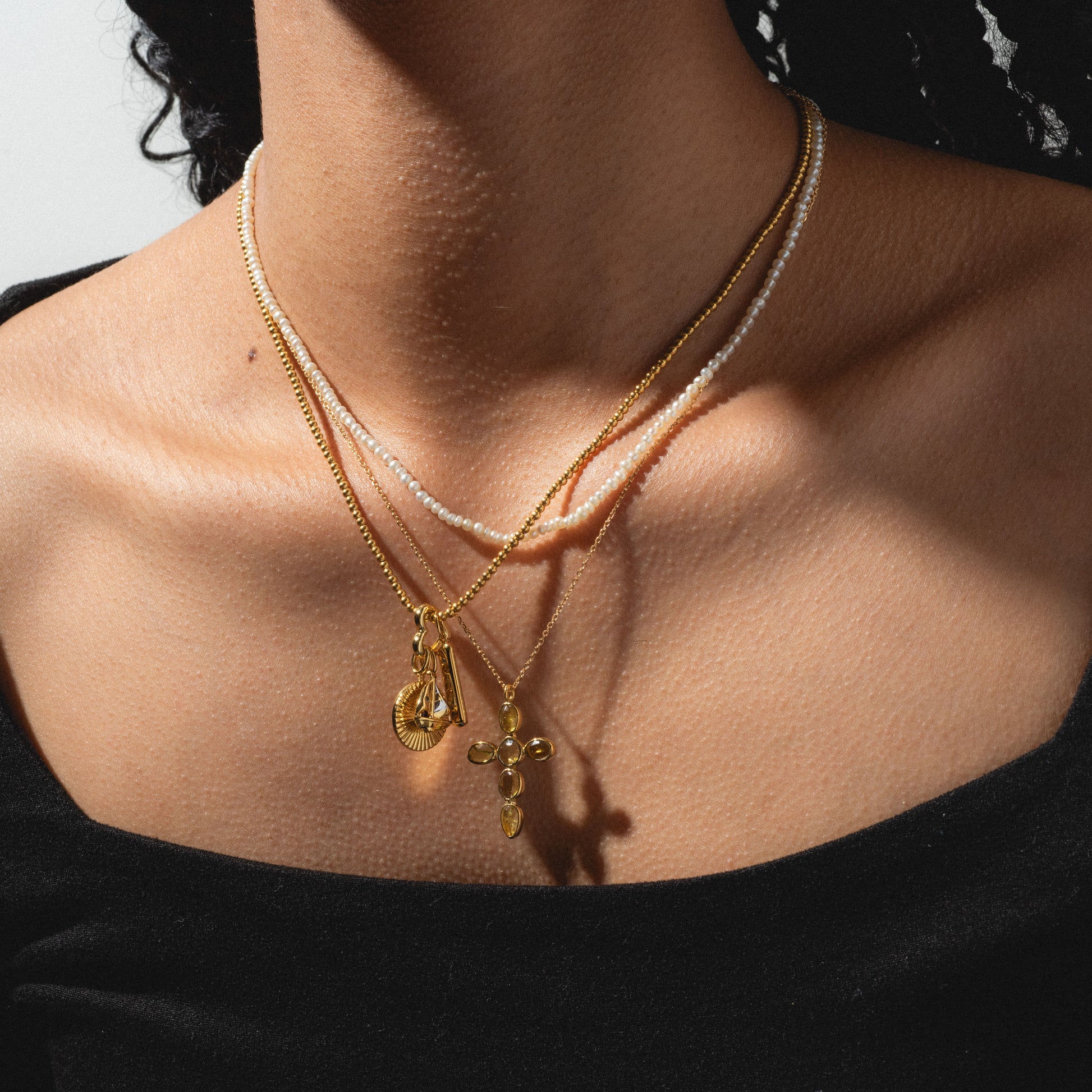 A close-up of a person in a black off-shoulder top wearing three layered necklaces: a white beaded strand, a gold chain with a Tiny Fuck Charm pendant, and another gold chain with a cross pendant.
