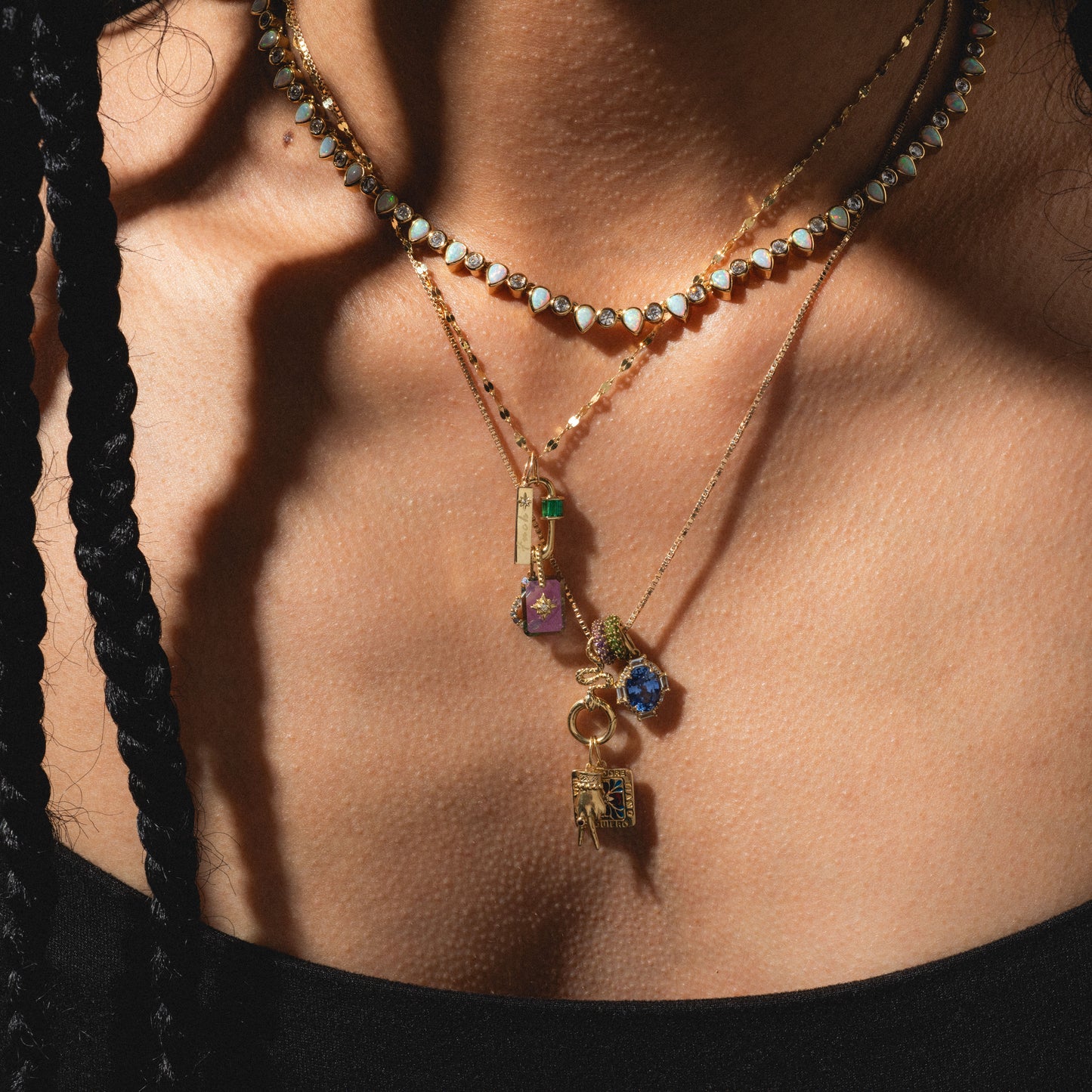 A close-up of a person in a black top, their braided hair and skin in natural light, wearing three layered gold necklaces including the Solid Gold Tiny Fuck Charm with gemstone and charm pendants.