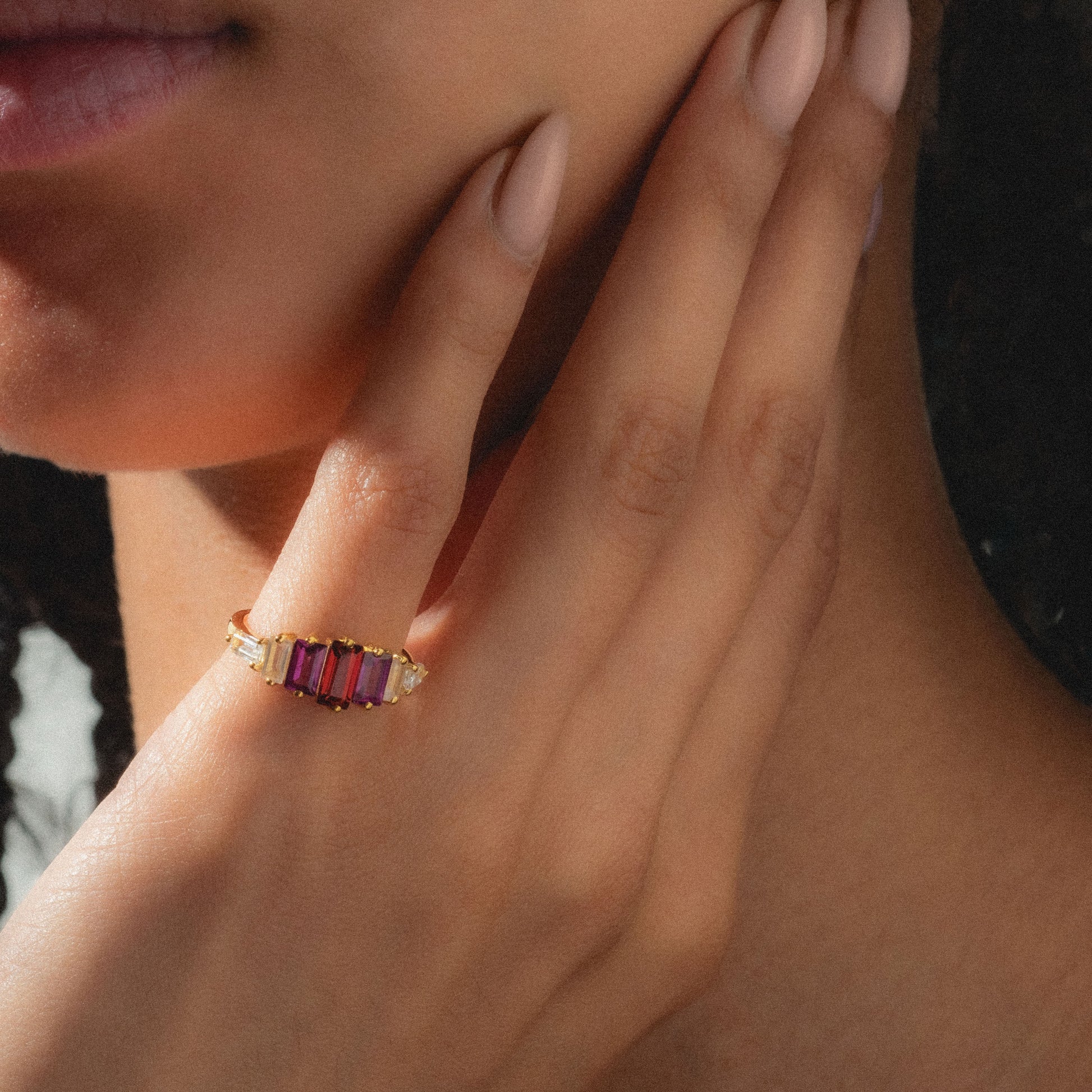 A close-up of a manicured hand touching the face highlights the Berry Baroque Garnet Crown Ring, featuring three rectangular red garnets set in gold vermeil. A softly blurred background enhances the ring's elegant, art deco-inspired design.