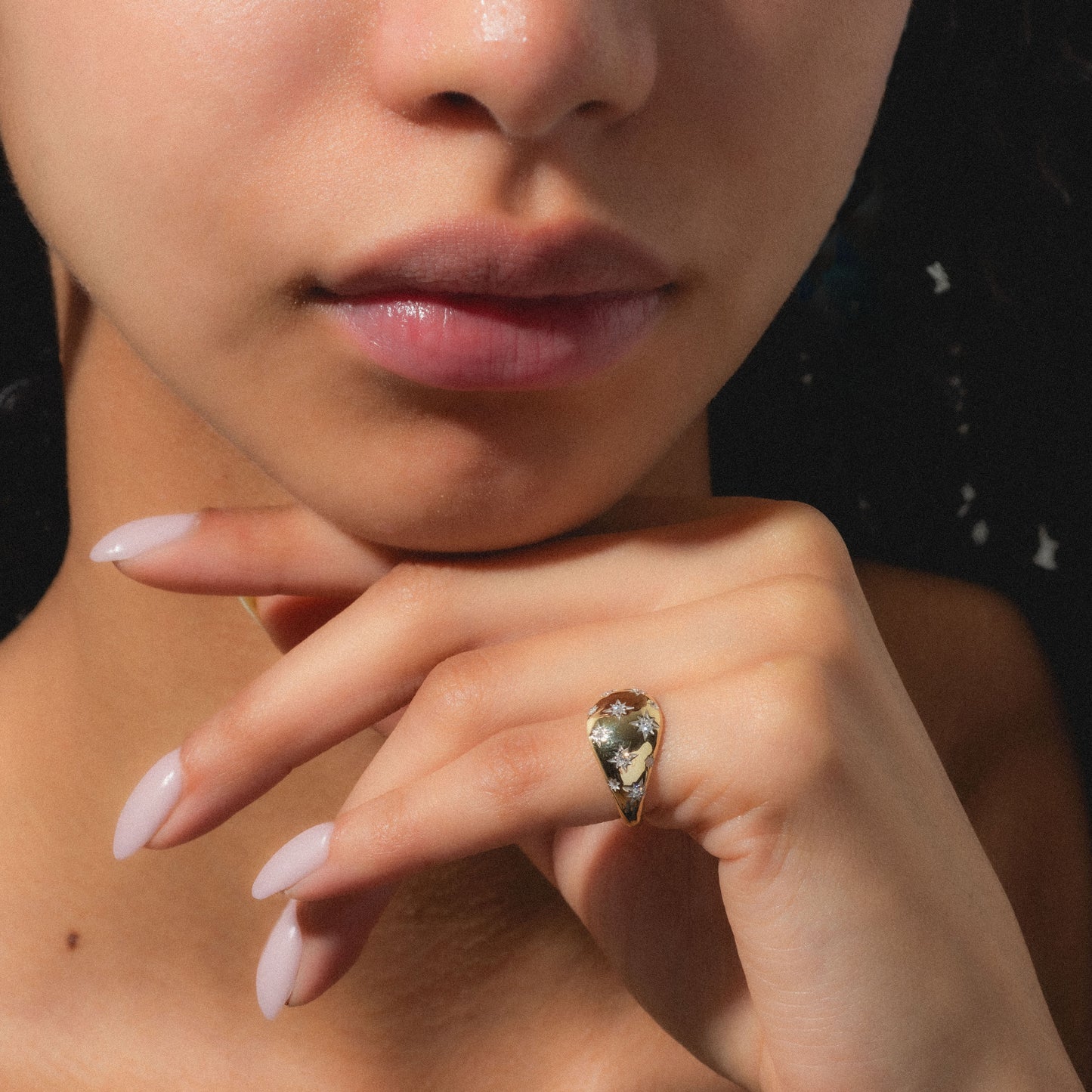 A person with smooth skin rests their chin on their hand, displaying the Solid Gold Star Crossed Dome Pinky Ring. The bold pinky ring stands out on manicured fingers with pale polish, as only the lower half of their face is shown.