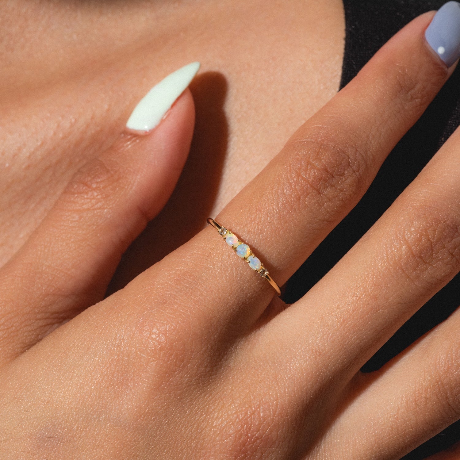 A hand with pastel-colored nails wears the Solid Gold 2025 October Capsule Classic Opal Trio Statement Ring, featuring three opals in a thin gold band, displayed against a soft, neutral background.
