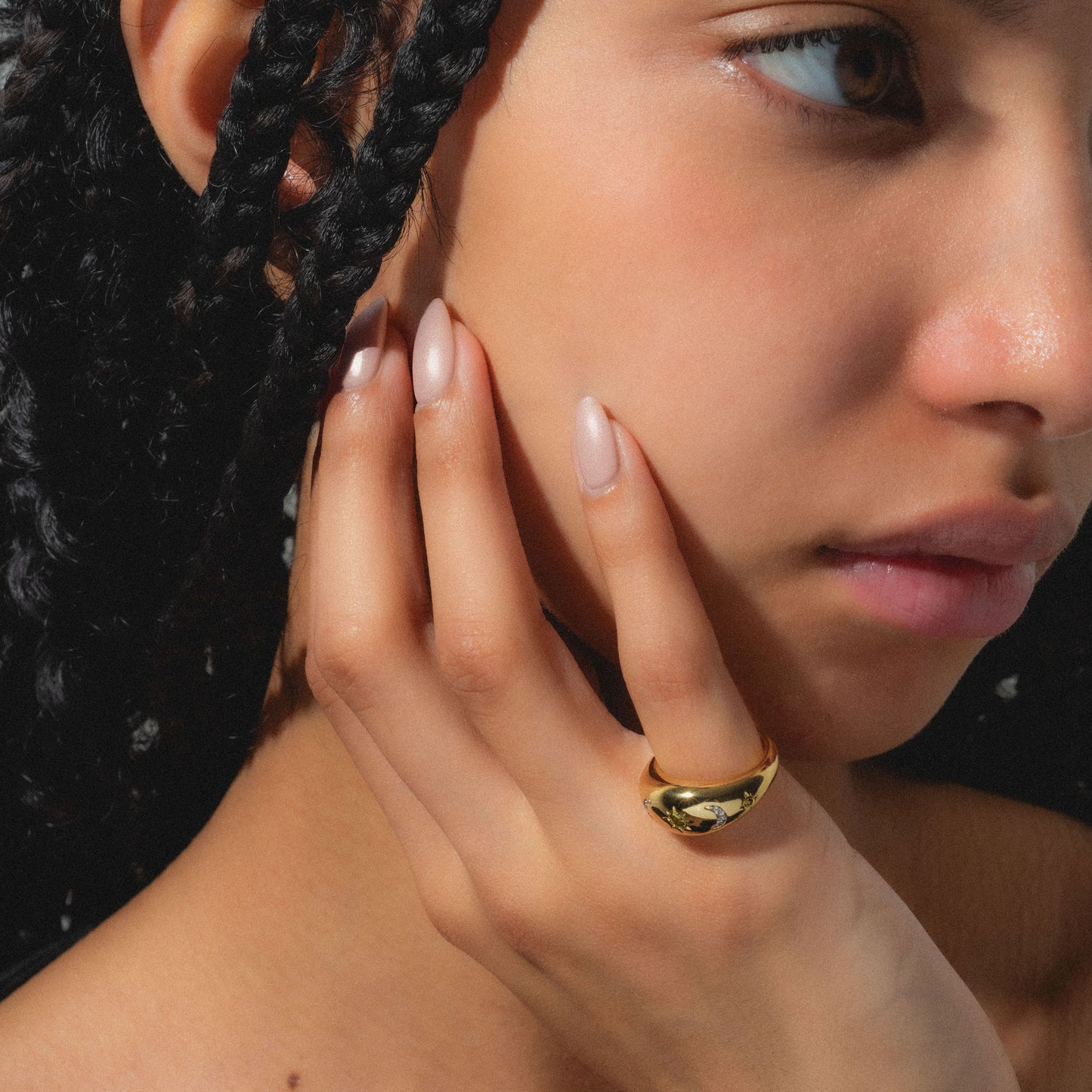 A close-up of a woman with braided hair and nude nails, showing natural makeup. She wears the Moonstruck Bold Dome Ring, a striking 14k gold plated piece that catches the light and highlights her smooth skin and jewelry.
