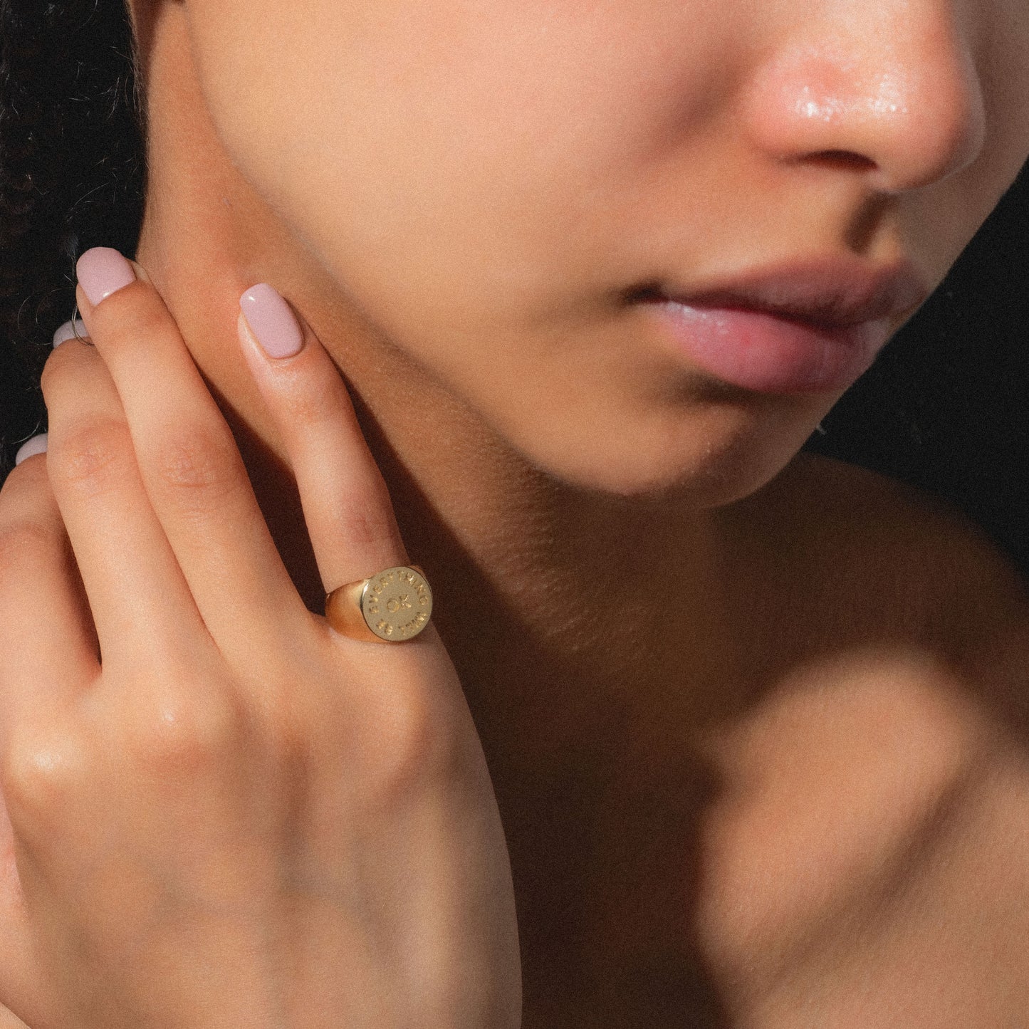 A person with smooth skin touches their neck, wearing the Everything Will Be Ok Pinky Signet Ring—14K gold plated and engraved with small symbols. Their lips are gently closed, and their nails are manicured in pale pink.