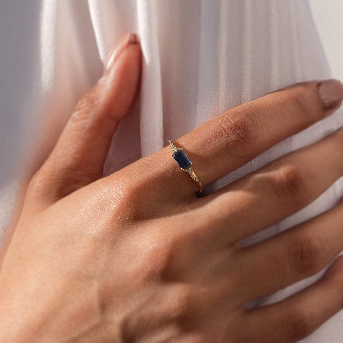 A close-up of a manicured hand on white fabric, showcasing the Solid Gold Glimmering Birthstone Baguette Ring set with a rectangular blue gemstone.