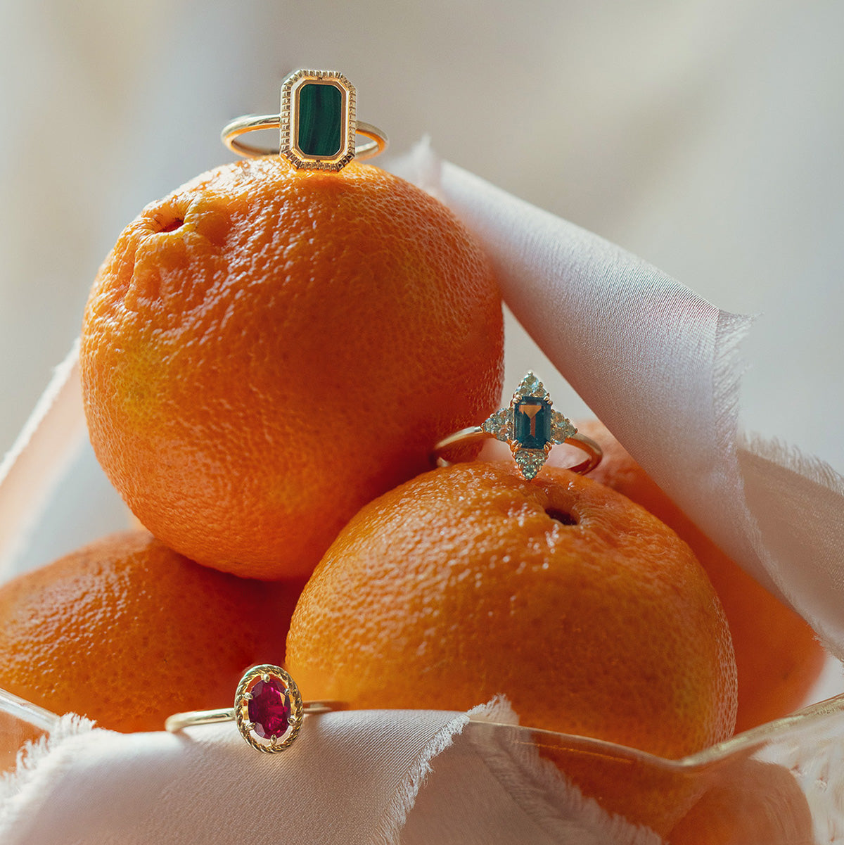 The 10k Gallery Frames Enamel and Malachite Reversible Ring shines atop bright oranges nestled in soft white cloth inside a glass bowl, its vibrant enamel and malachite contrasting beautifully with the fruit.