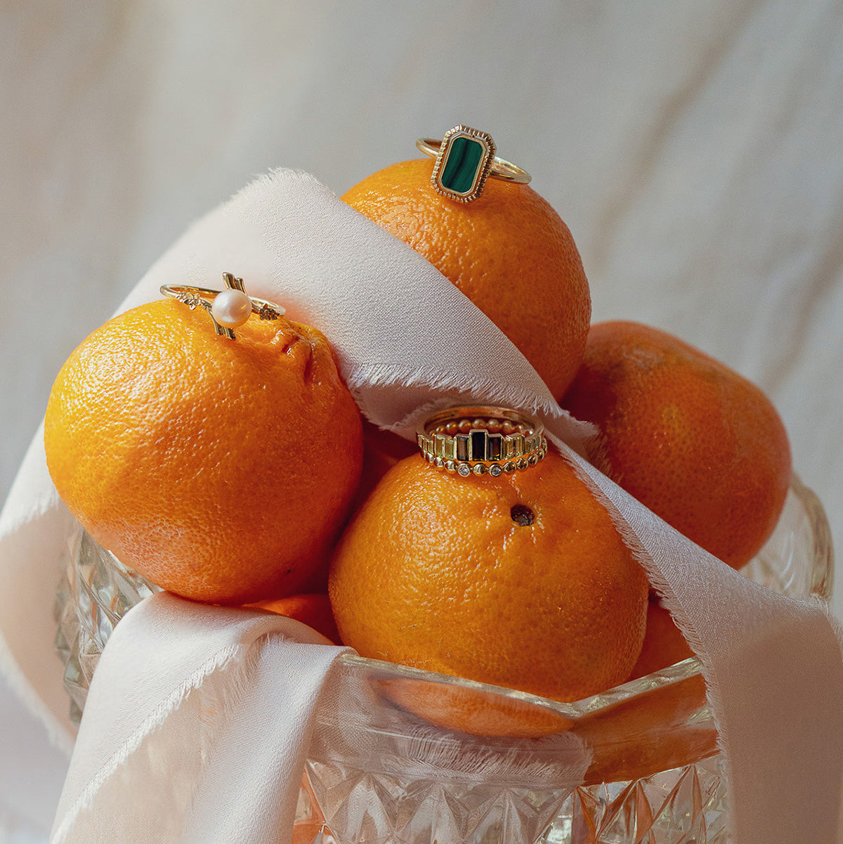 The 10k Green Tourmaline Skyline Band is displayed atop oranges in a glass bowl, accented by a light silky ribbon draped over the fruit, with a softly blurred background.