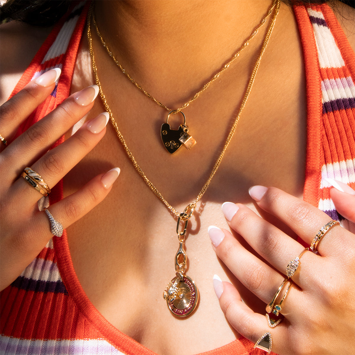 A woman wearing Charms by Mod + Jo layered gold necklaces and plated charms, with gemstone rings, is shown from the shoulders down in an orange and white striped deep-neckline top. Her nails are manicured a soft pink.