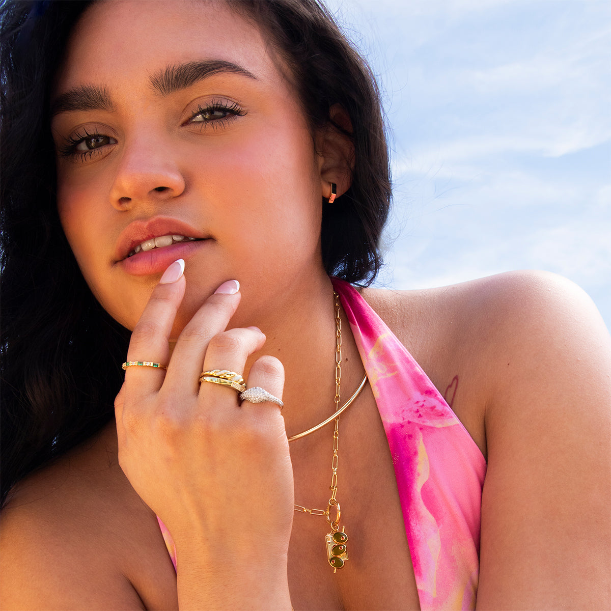 A woman with long dark hair poses outdoors against a blue sky, wearing a pink patterned halter top and layered gold jewelry, including the Ari Hoops earrings. She touches her lips and chin thoughtfully.