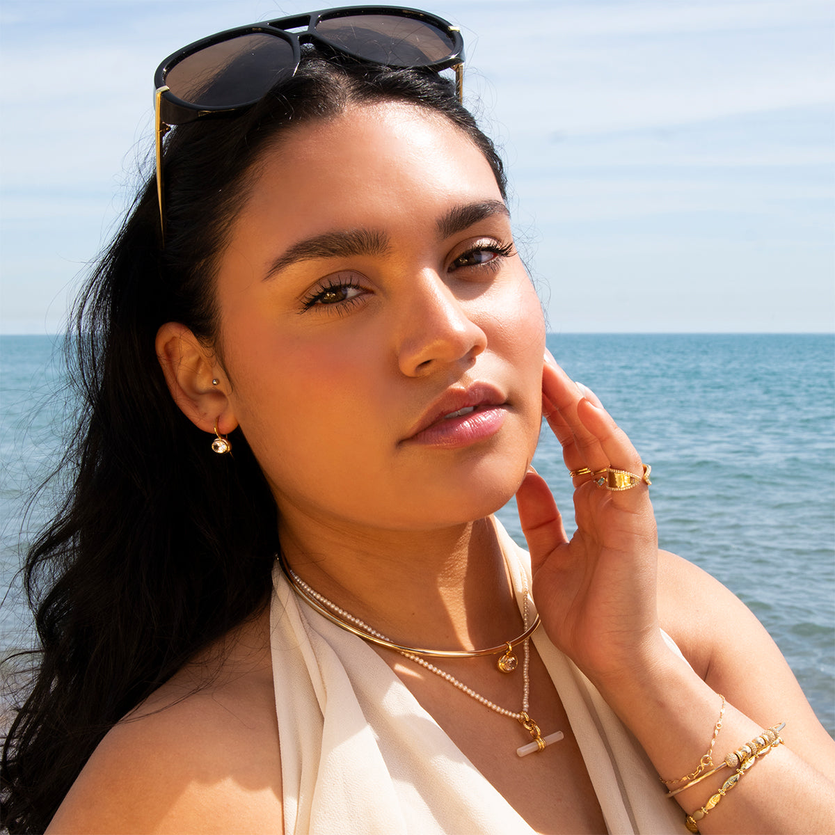 A woman with long dark hair and sunglasses on her head wears Lucia Oval French Hook Earrings and a white halter top, standing by the ocean under a blue sky, touching her face and looking at the camera.