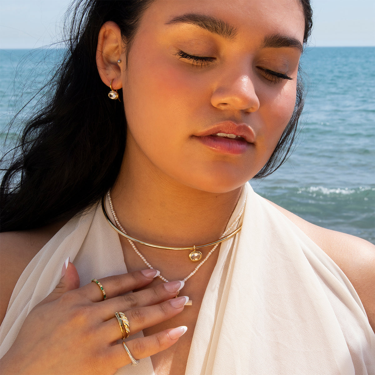 A woman with long dark hair stands by the sea, eyes closed, wearing Lucia Oval French Hook Earrings along with layered gold jewelry and rings, paired with a sheer white top and natural makeup.