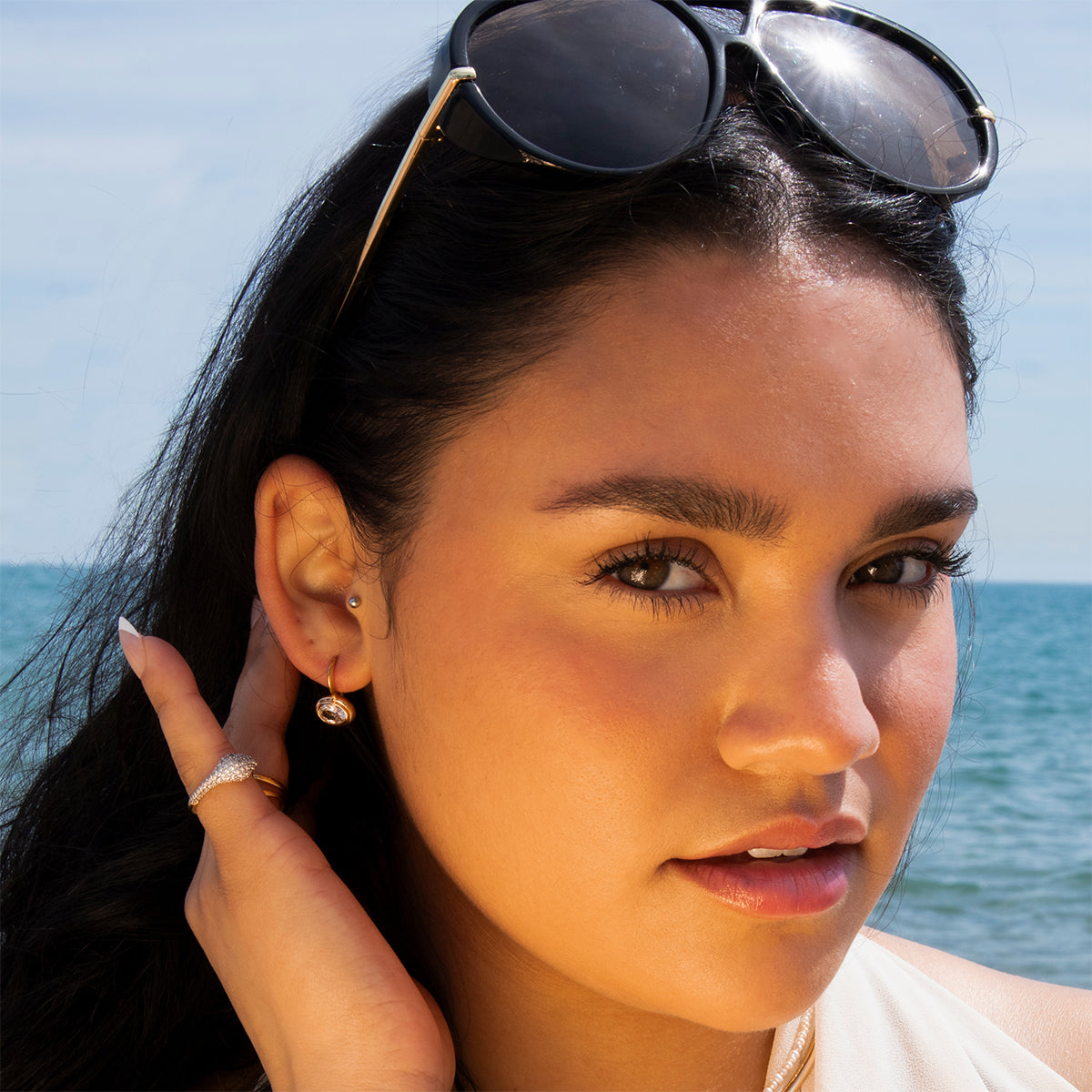 A woman with long dark hair wears Lucia Oval French Hook Earrings, sunglasses on her head, and a ring as she poses by the ocean under bright sunlight, touching her ear and looking at the camera.
