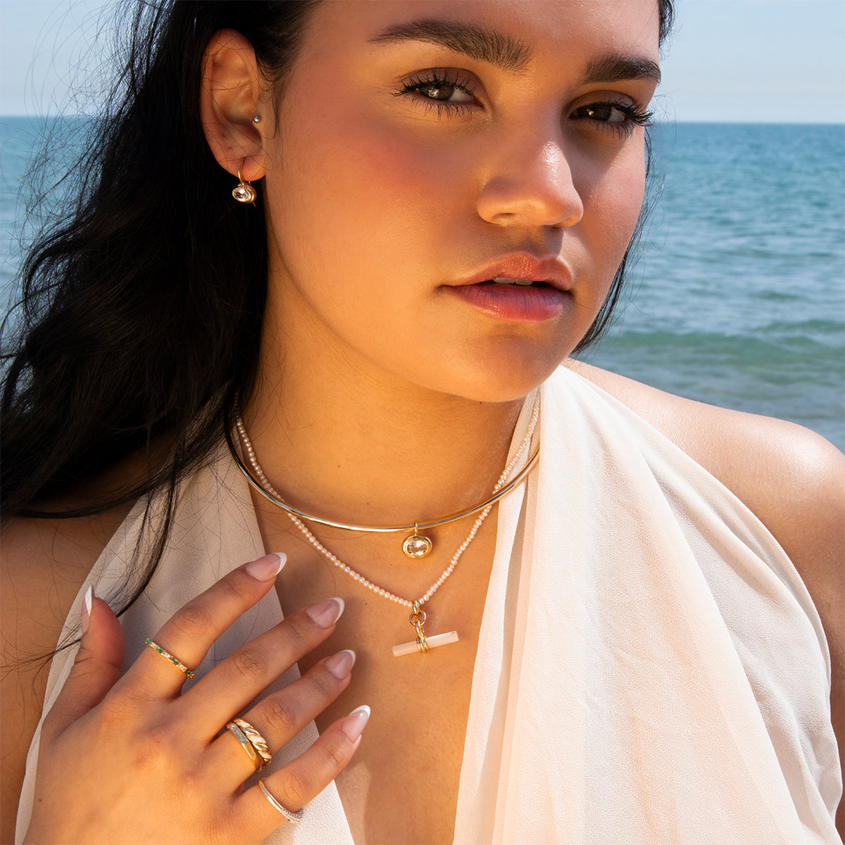 A woman with long dark hair stands by the ocean in a white sleeveless top, gently touching her Hayes Collar Necklace in 18k yellow gold. Layered gold jewelry and her calm expression match the minimal, serene seaside setting.