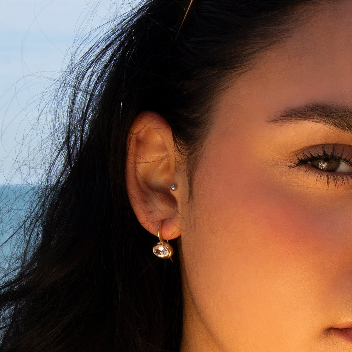 Close-up of a woman's face and ear wearing the Lucia Oval French Hook Earrings. The ocean and sky appear blurred in the background.