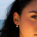 Close-up of a woman's face and ear wearing the Lucia Oval French Hook Earrings. The ocean and sky appear blurred in the background.