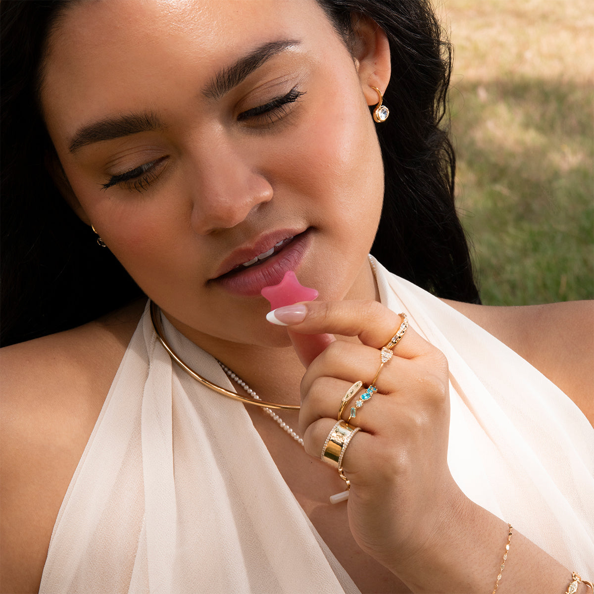 A woman with long dark hair and glowing skin, wearing a white halter dress and Lucia Oval French Hook Earrings, holds a pink star-shaped gummy near her lips against a grassy background.