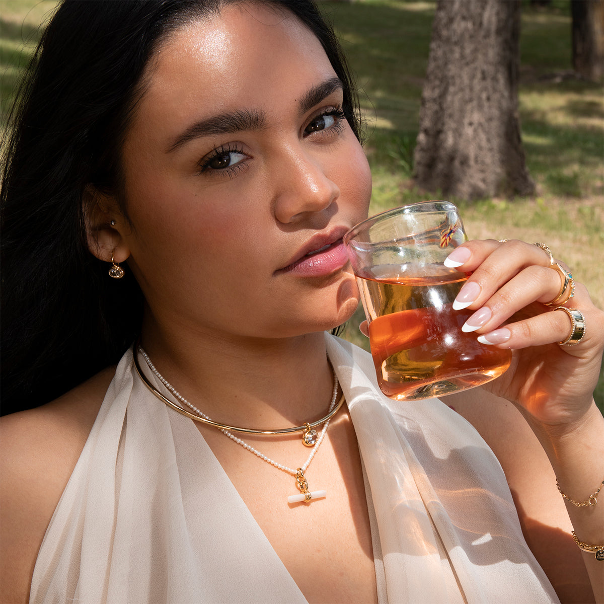 A woman with long dark hair wears the minimalist Hayes Collar Necklace in 18k yellow gold while holding an amber drink close to her lips, seated outdoors on sunlit grass with trees in the background.