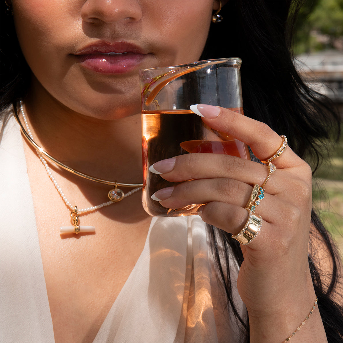 A woman with manicured nails holds a golden drink in a glass beaker. She wears layered necklaces, including the Hayes Collar Necklace, and multiple rings, with sunlight highlighting her minimalist jewelry and outfit.