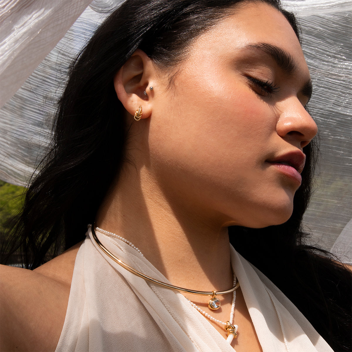A woman with long dark hair closes her eyes peacefully, wearing Levi Huggies gold earrings and pearl jewelry. Sunlight highlights her glowing skin against a sheer white fabric backdrop as she dons a light, draped top.