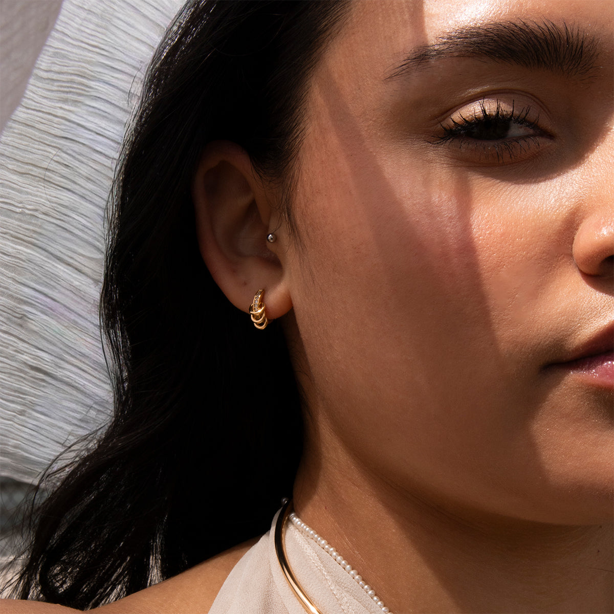 Close-up of a woman's face in sunlight, highlighting her smooth skin and dark hair. Her ear is adorned with the Levi Huggies, small gold hoop earrings, and a stud, with shadows and soft light enhancing the details.