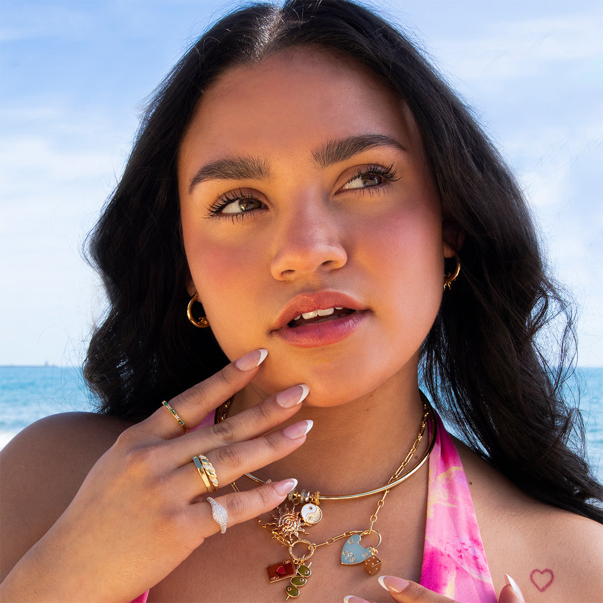 A woman with long dark hair stands on a beach, wearing Cherub Hoops and layered gold necklaces. She has manicured nails, a small heart tattoo on her shoulder, and is dressed in a pink top, gazing thoughtfully into the distance.