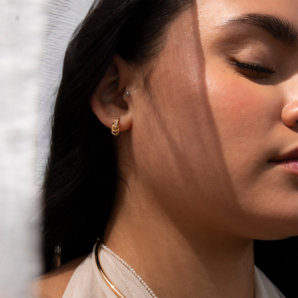 A close-up of a woman with closed eyes wearing Levi Huggies gold hoop earrings and a pearl necklace, sunlight and soft shadows across her face highlighting her skin's warm, natural glow.