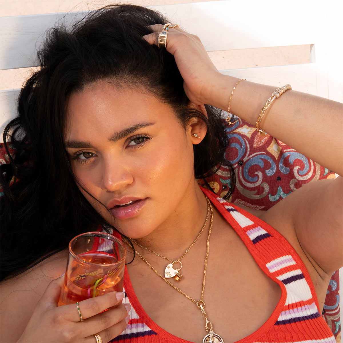 A woman with long dark hair lounges on a cushion, wearing a striped red, white, and pink top and Charms by Mod + Jo layered as necklaces, bracelets, and rings while holding a citrus drink and looking at the camera.