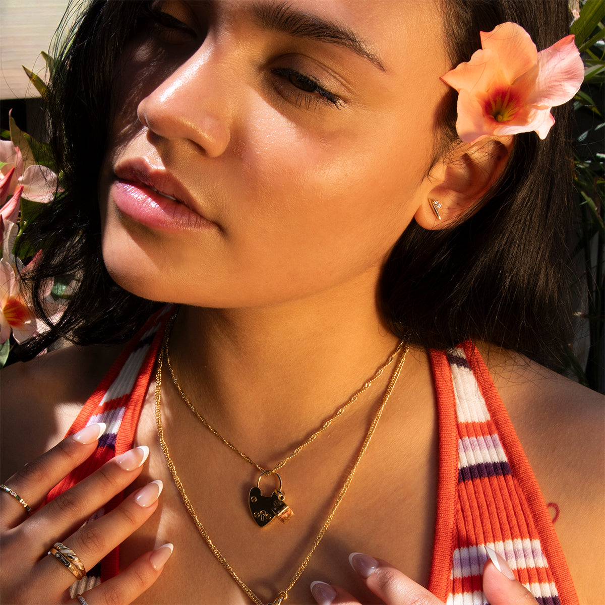 A woman with smooth skin and a peach blossom in her hair stands in sunlight among flowers, wearing layered gold necklaces—including a heart-shaped locket—a striped orange and white top, and the Ida Bar Studs.