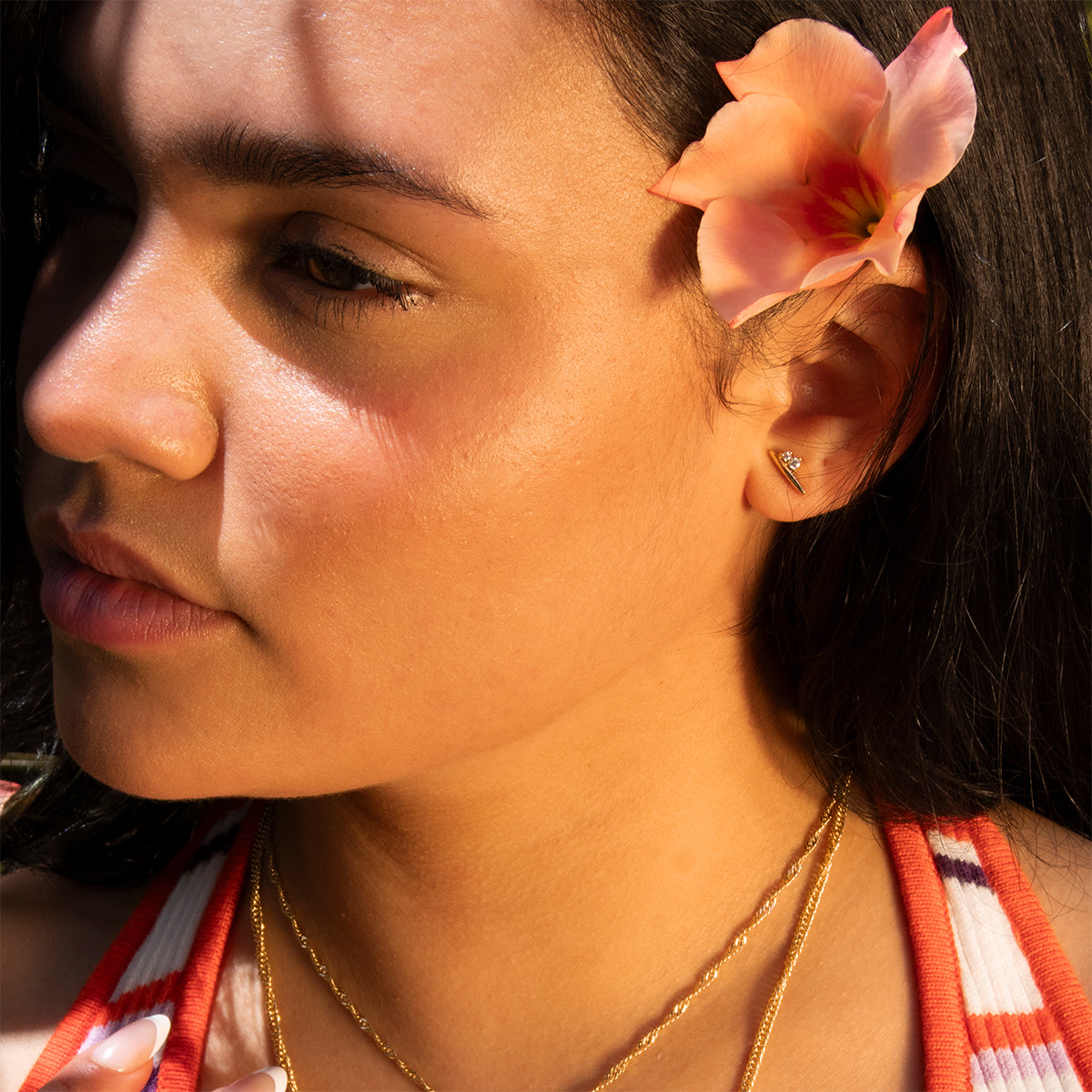 A woman with long dark hair wears a peach flower behind her ear, the Ida Bar Studs, layered gold necklaces, and a striped top. Sunlight and shadows fall across her face as she looks to the side.
