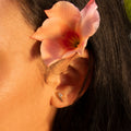 Close-up of a person with long dark hair, wearing gold Ida Bar Studs featuring a small gemstone. An orange-pink flower is tucked behind the ear. Sunlit skin and blurred background complete the scene.