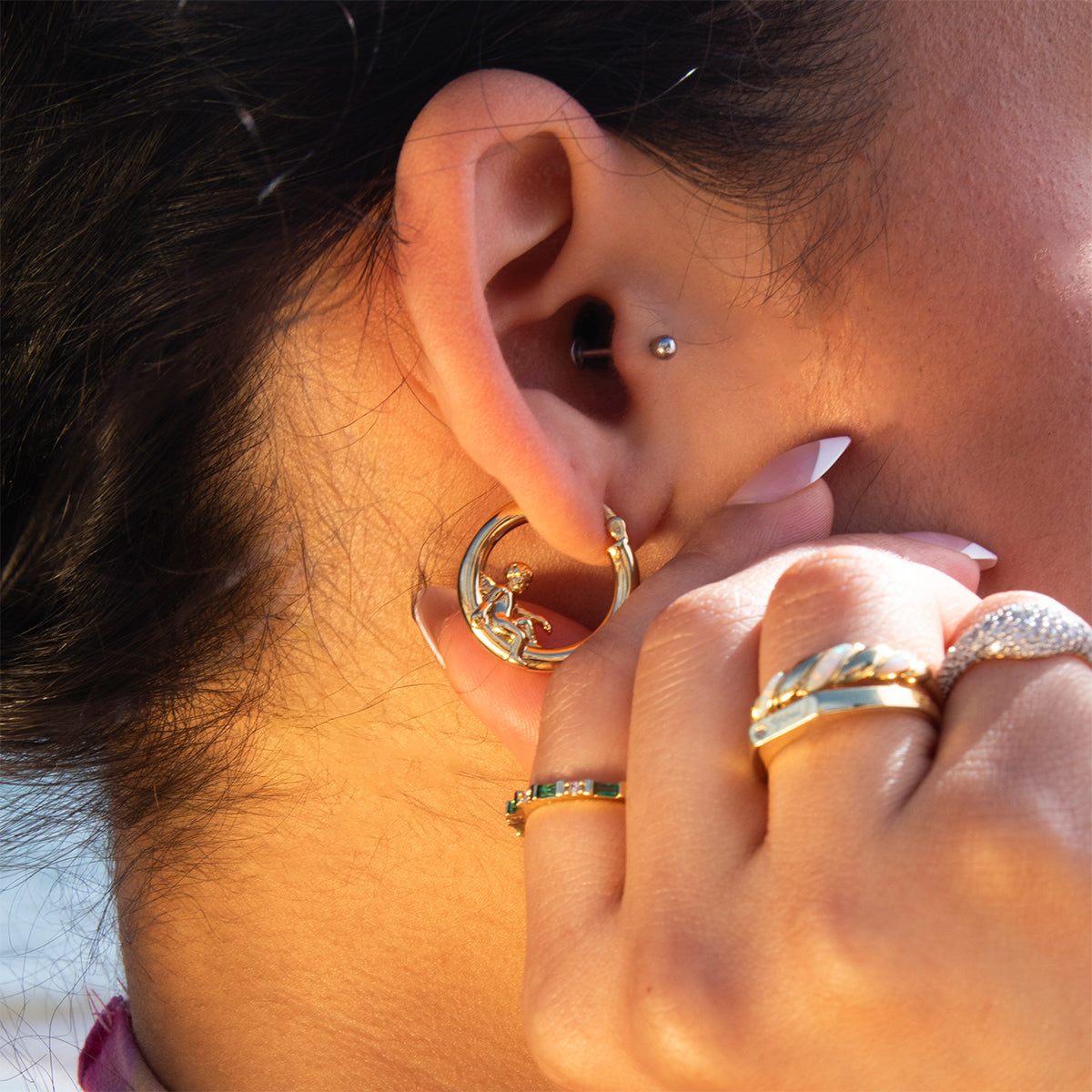 Close-up of a woman's ear with multiple piercings, featuring Cherub Hoops gold earrings and a small stud. Her hand, adorned with rings and manicured nails, rests near her ear.