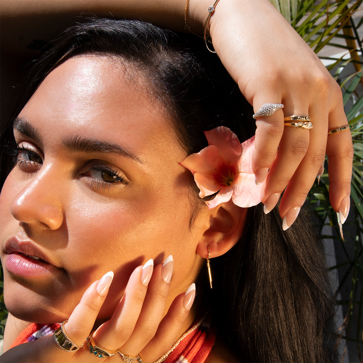 A woman with smooth skin poses in sunlight, holding a pink flower near her ear. She wears Mila Threader Earrings along with gold rings and bracelets. Her manicured nails stand out as dark hair frames her face, with green leaves in the background.