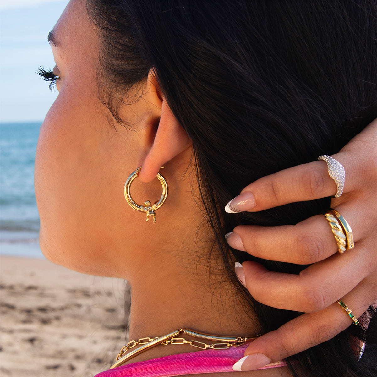 A woman with long dark hair poses by the beach in a pink top, showcasing Cherub Hoops gold earrings, layered gold necklaces, and intricately designed rings as her hand rests near her ear.