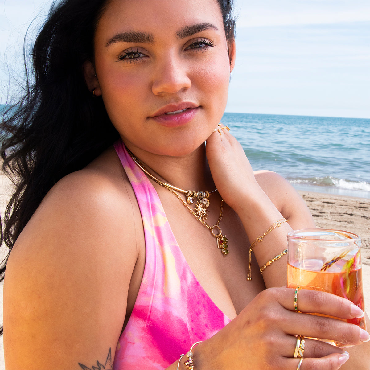 A woman wearing the Hayes Collar Necklace and a pink tie-dye swimsuit holds a fruity drink while standing on a sandy beach, with the ocean and blue sky in the background.