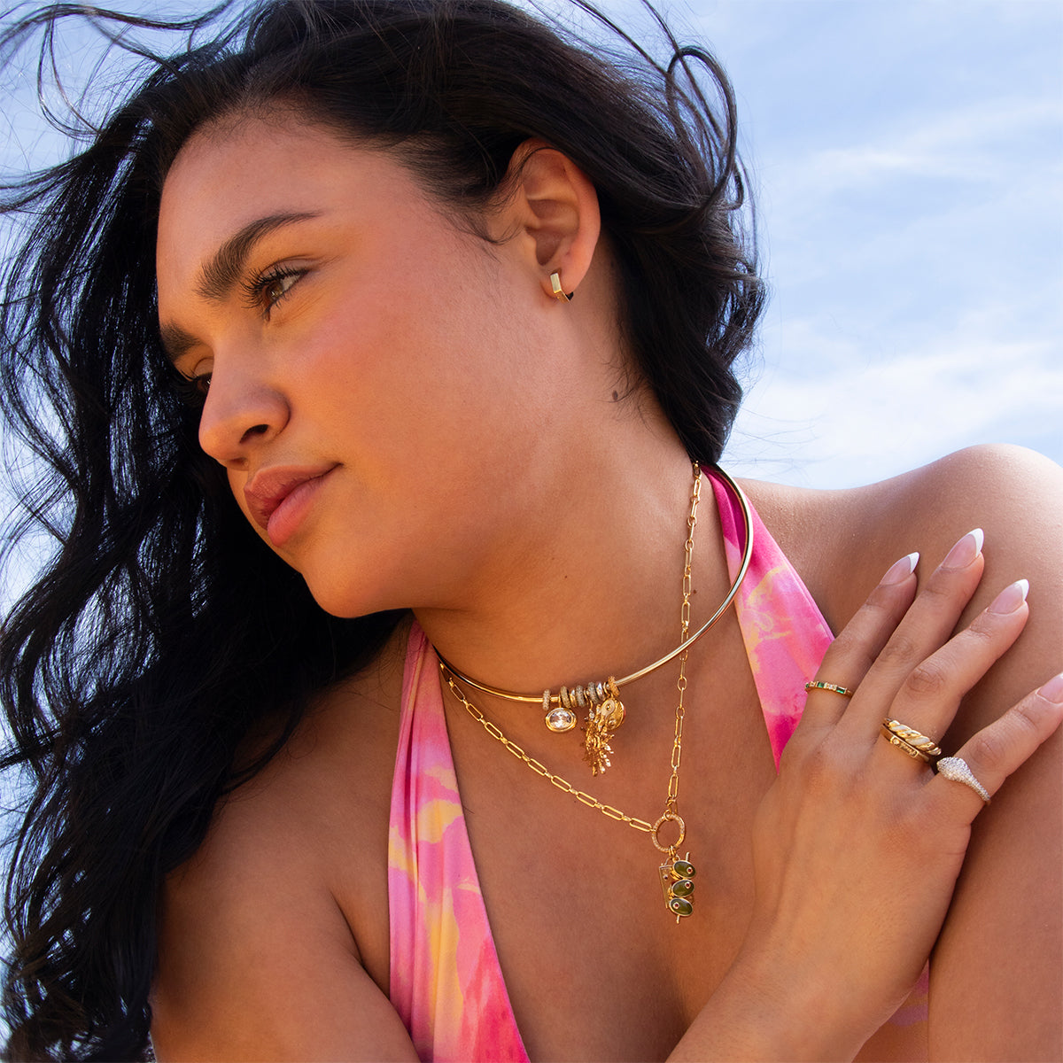 A woman with long dark hair wears gold jewelry, including Ari Hoops earrings, layered necklaces, and rings. She is dressed in a pink and orange patterned top while posing outdoors against a blue sky.