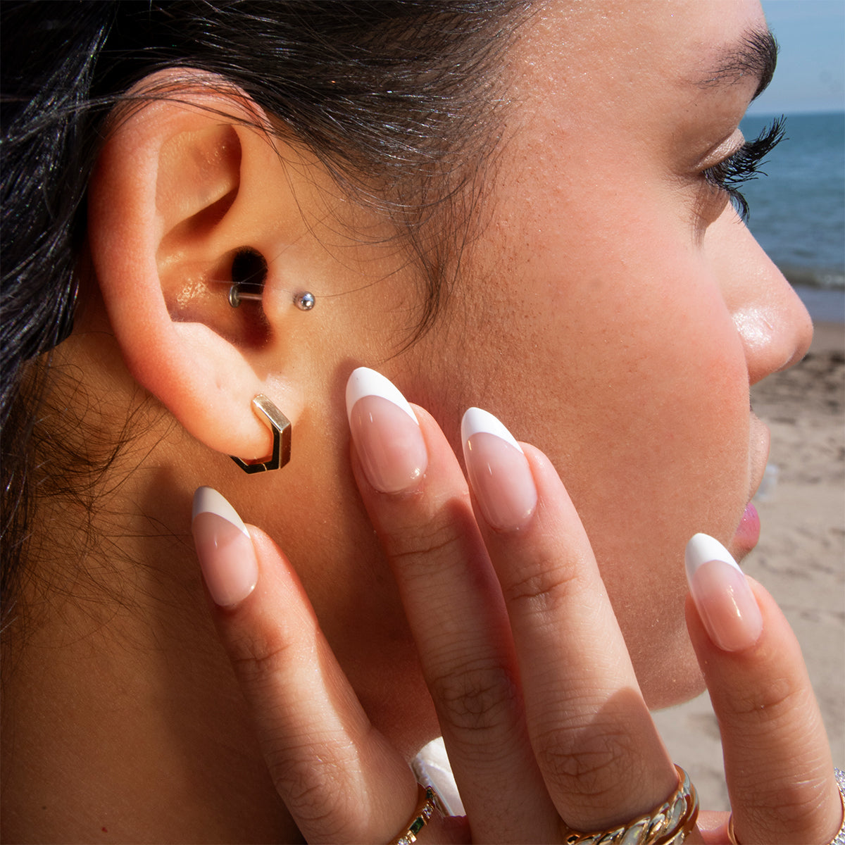 A close-up of a person's ear adorned with Ari Hoops and other gold earrings. Their hand with French-tipped nails and gold rings touches their face, set against a sandy beach and ocean backdrop.
