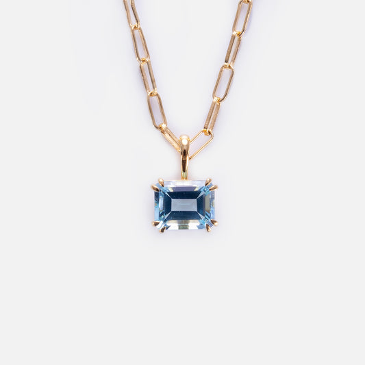 The Born to Shine Birthstone Necklace showcases a gold paperclip chain with elongated links and a rectangular, faceted light blue gemstone pendant in a gold prong setting, displayed on a plain white background.