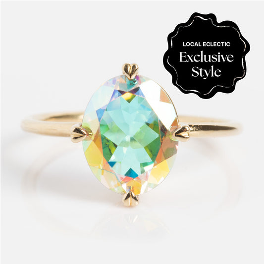 The One of a Kind 14k Heart Prong Angel Aura Ring features an oval iridescent gemstone held by heart-shaped prongs. A black label in the corner states, Local Eclectic Exclusive Style.