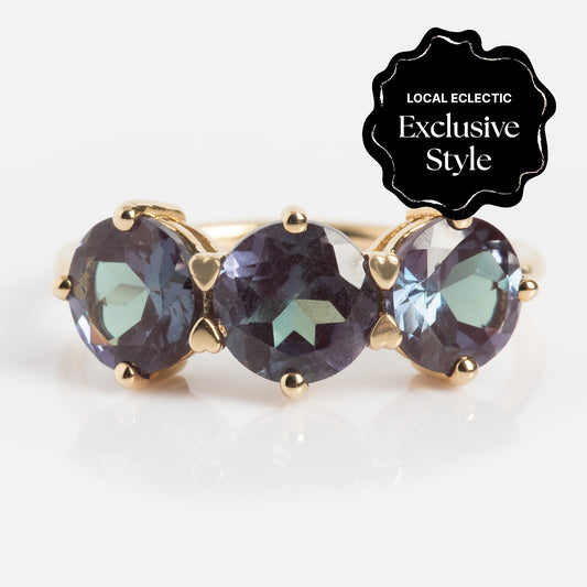 The One of a Kind 14k Heart Prong Lab Alexandrite Trio Ring features three round, purple-green iridescent stones set side by side. A black label in the corner reads Local Eclectic Exclusive Style.