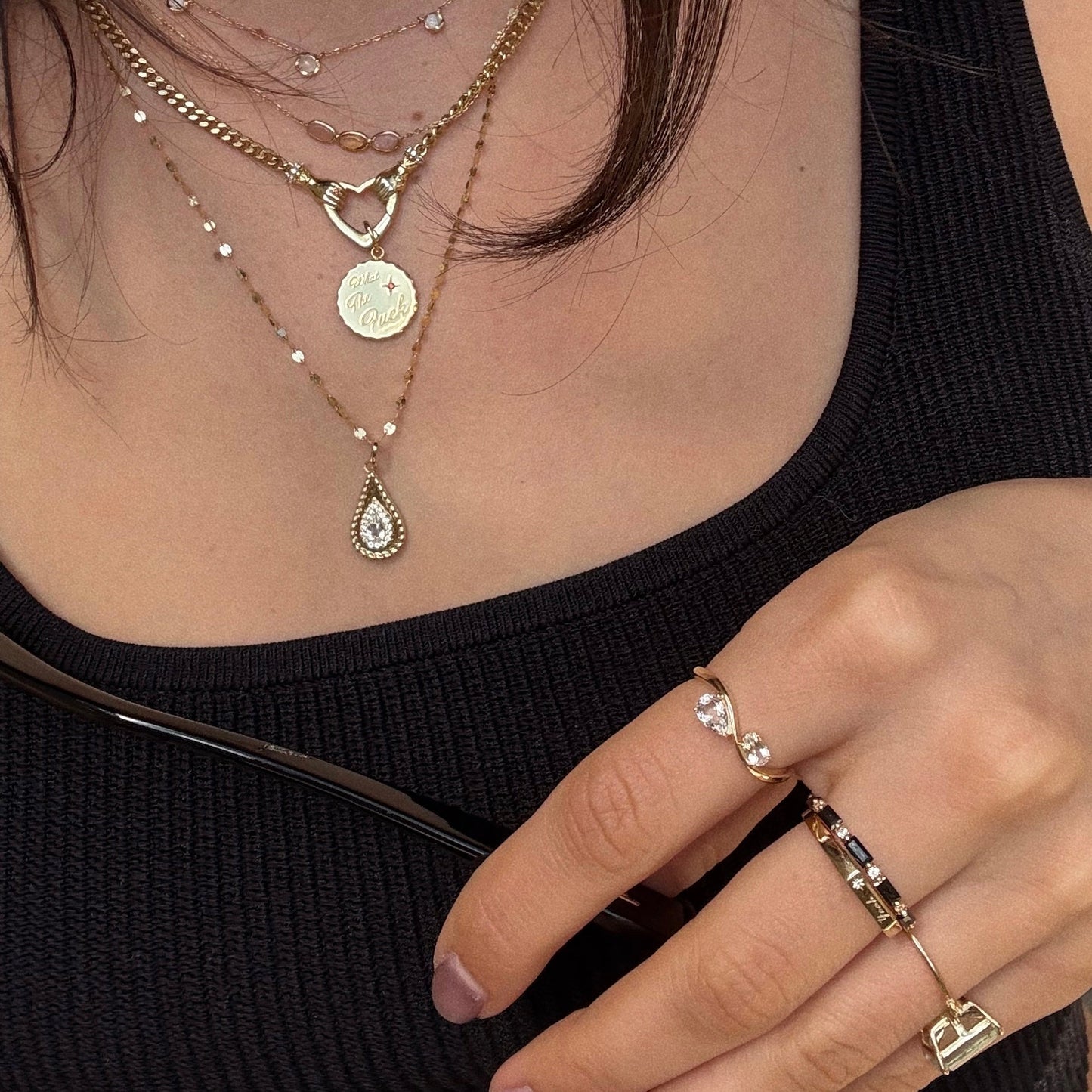 A close-up of a person in a black ribbed tank top wearing the Solid Gold 2026 April White Topaz Capsule Set—layered gold necklaces with a heart locket, teardrop white topaz pendant, and gold rings with stones—with free gift.