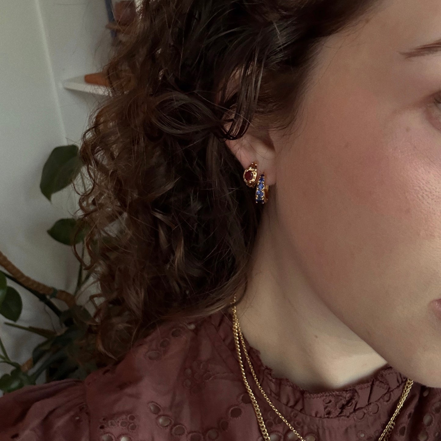 A person with curly brown hair wears a maroon ruffled top and layered gold necklaces, accentuated by Gilded Garnet Huggie Hoops. A green leafy plant can be seen in the background.