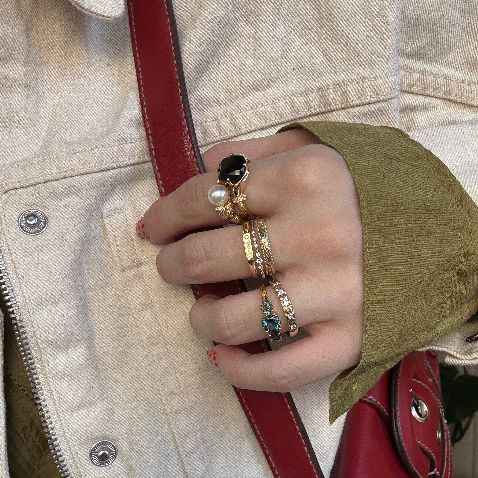 A hand wearing multiple gold rings, including the Pearl Siren Statement Ring, holds a red handbag strap. The wearer has on a cream jacket layered over a green sleeve.