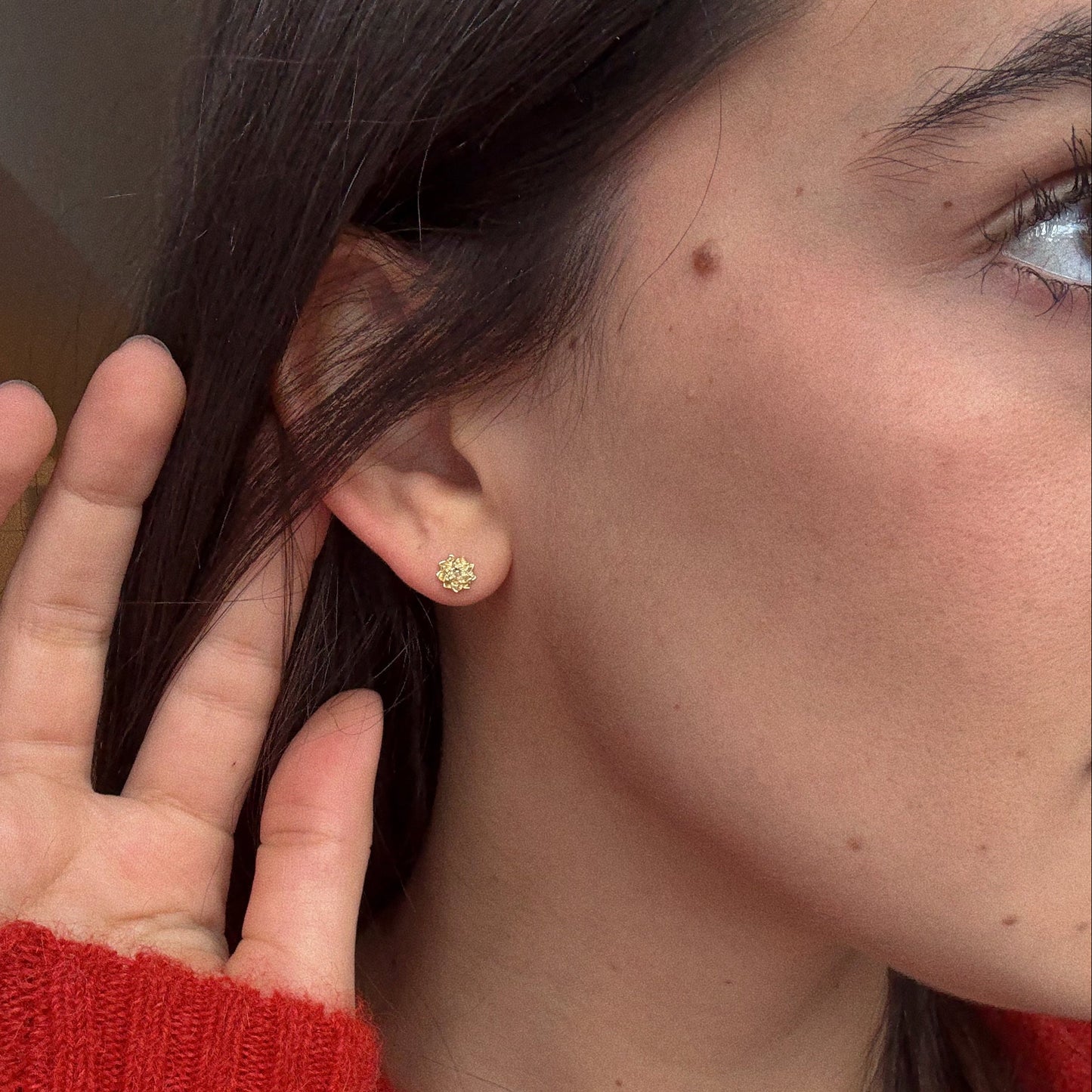 A close-up shows a person wearing Solid Gold Birth Flower Studs—a small, gold, flower-shaped earring—while holding their hand near their ear, dark hair framing their face and a red sweater adding a cozy touch.