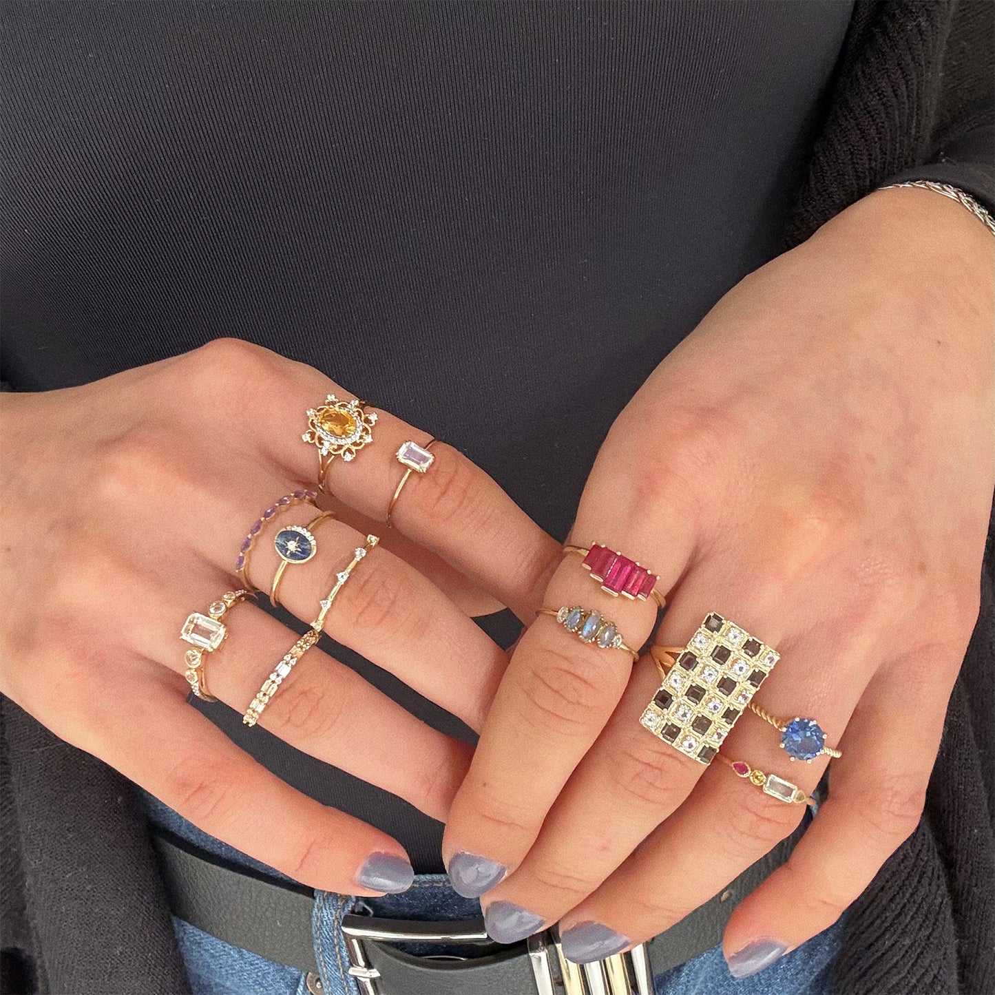 A person with painted nails showcases both hands adorned with Solid Gold Surprise Rings and gemstone rings featuring sapphires, diamonds, and rubies, over a dark top and black cardigan.