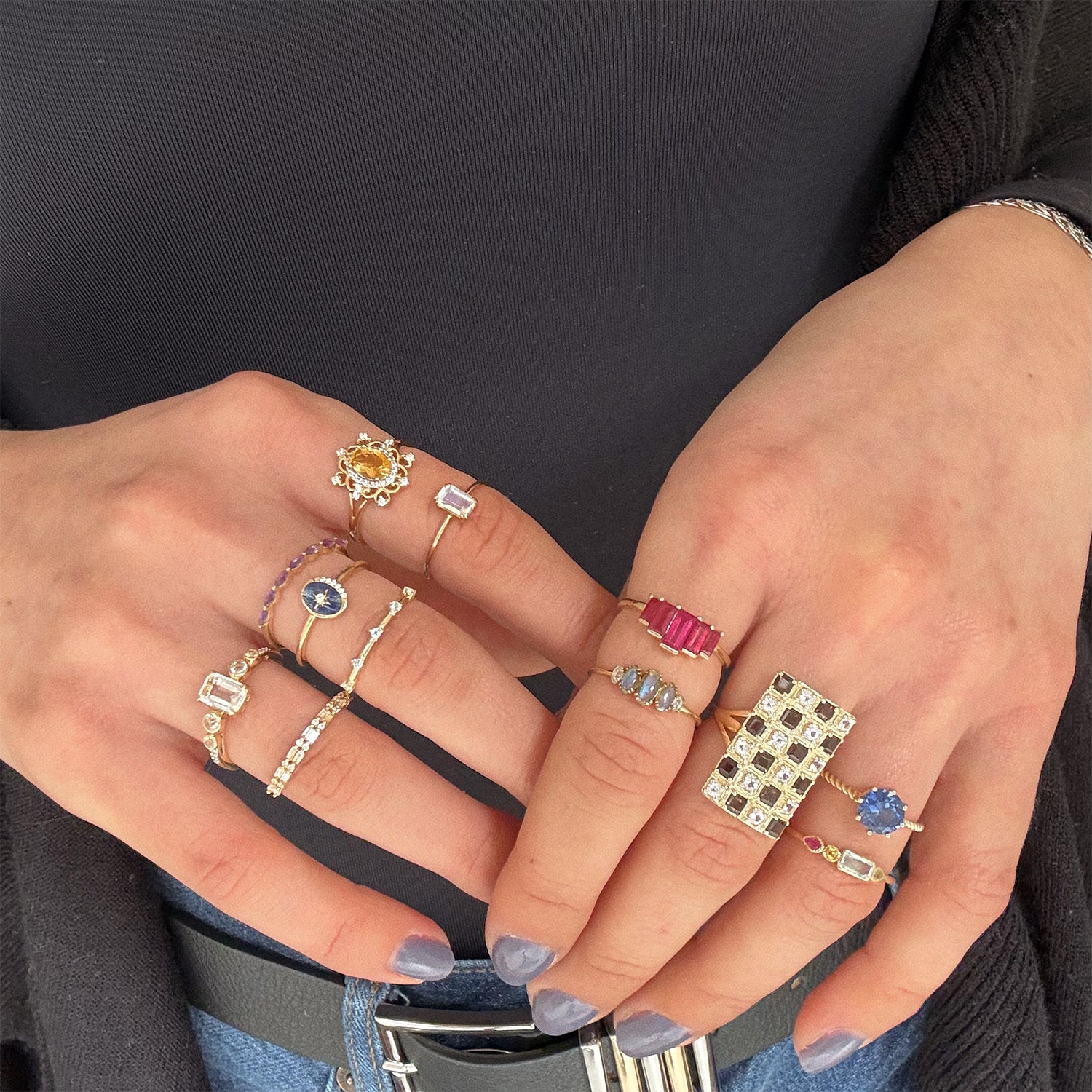 A person with painted nails showcases both hands adorned with Solid Gold Surprise Rings and gemstone rings featuring sapphires, diamonds, and rubies, over a dark top and black cardigan.