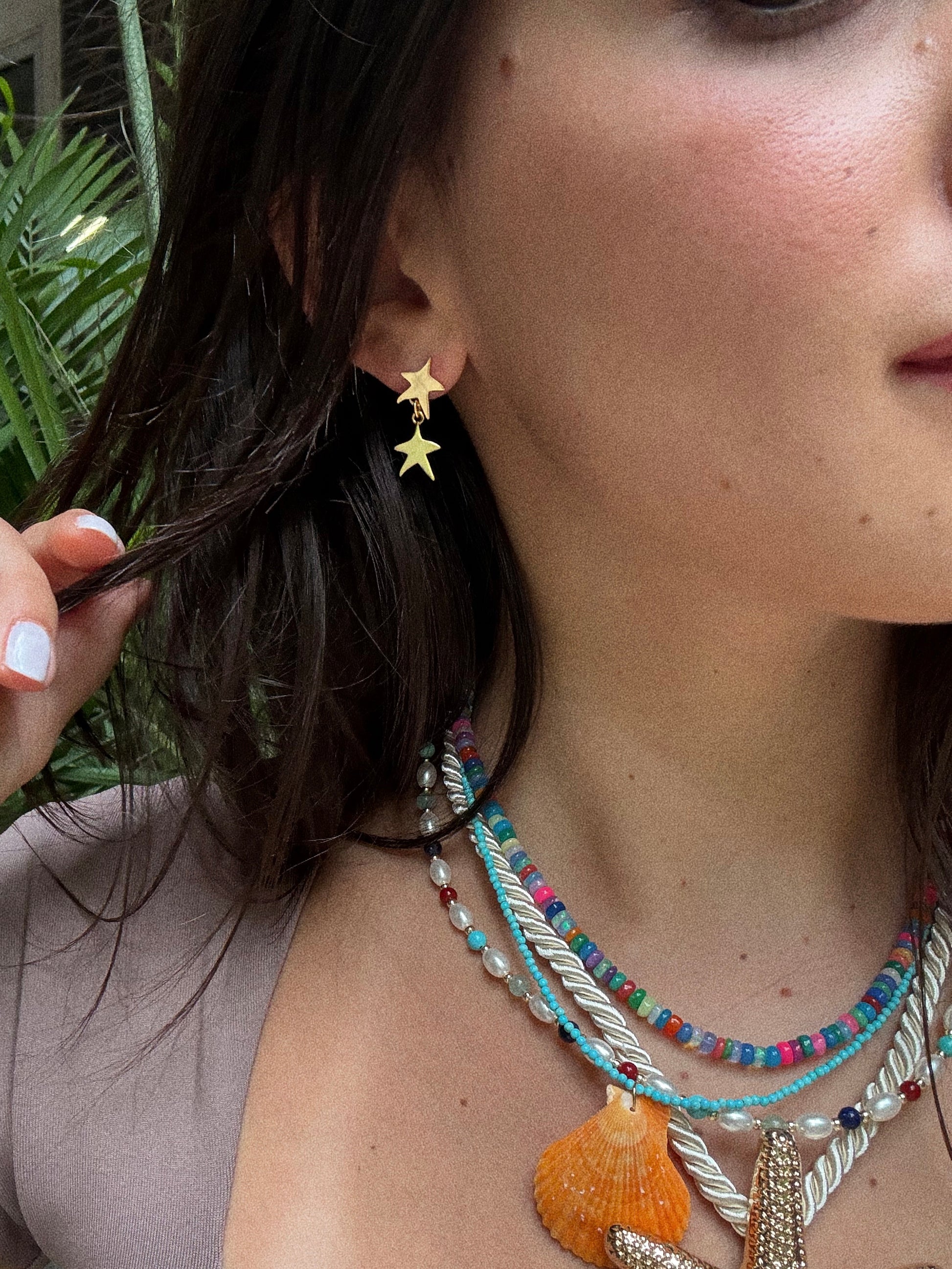 A woman wearing Star Drop Earrings and colorful beaded necklaces, one featuring a large orange seashell pendant, holds a strand of her brown hair with green plants in the background.