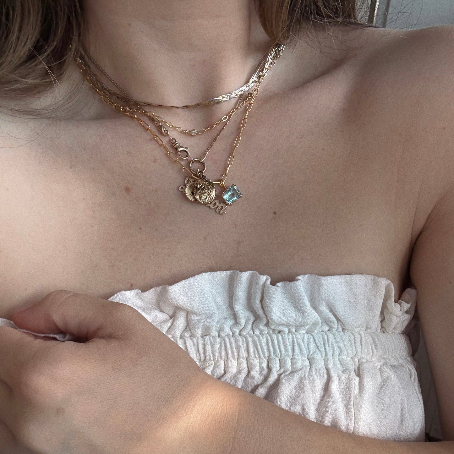 A person wears layered gold necklaces, including a heart charm and the Born to Shine Birthstone Necklace with a blue stone, styled with a white, strapless ruffled top. Their hand rests on the top’s edge near their shoulder.