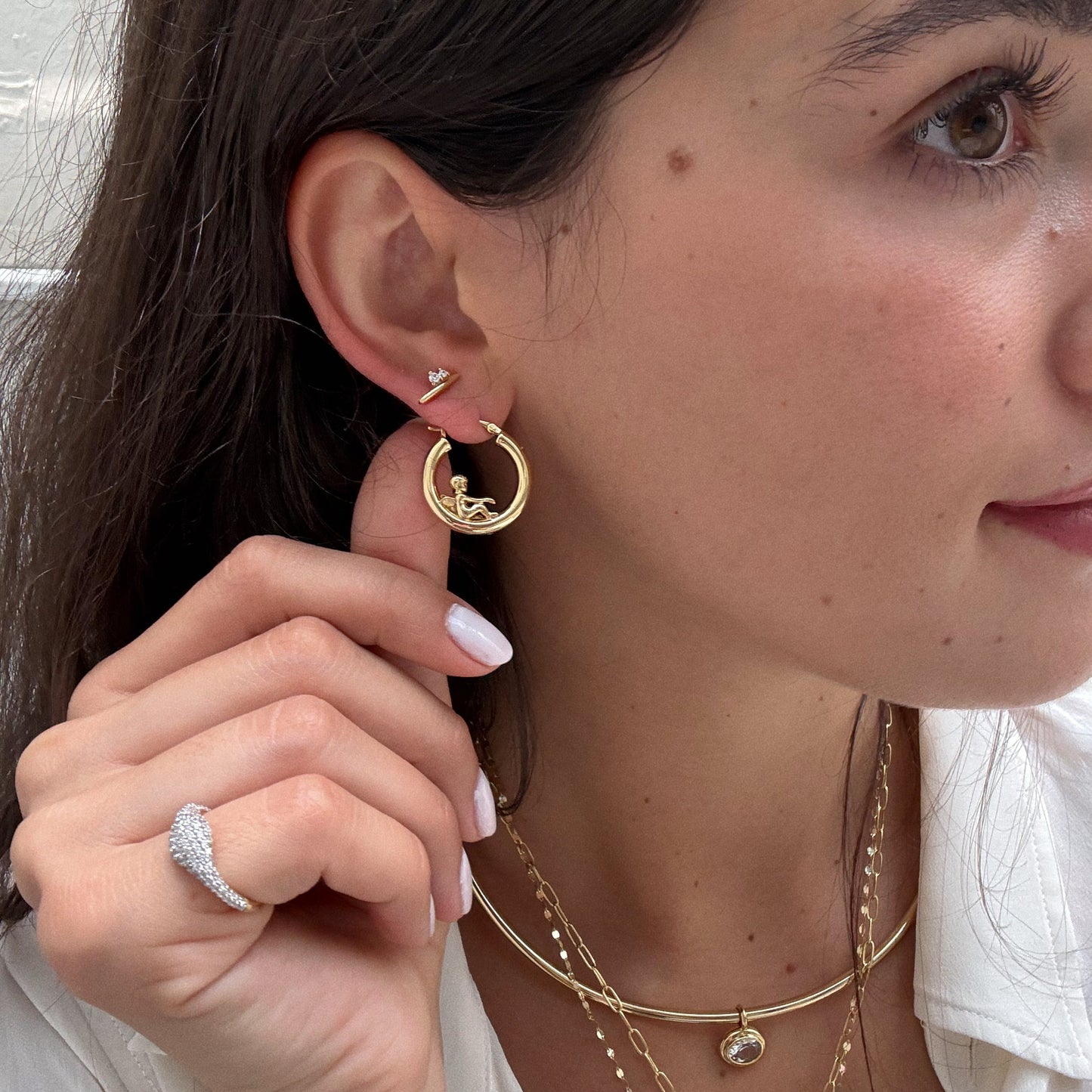 A woman with long brown hair and pale pink nails wears gold jewelry, including Ida Bar Studs, layered necklaces, a diamond ring, and holds a large hoop earring. She is dressed in a white top.