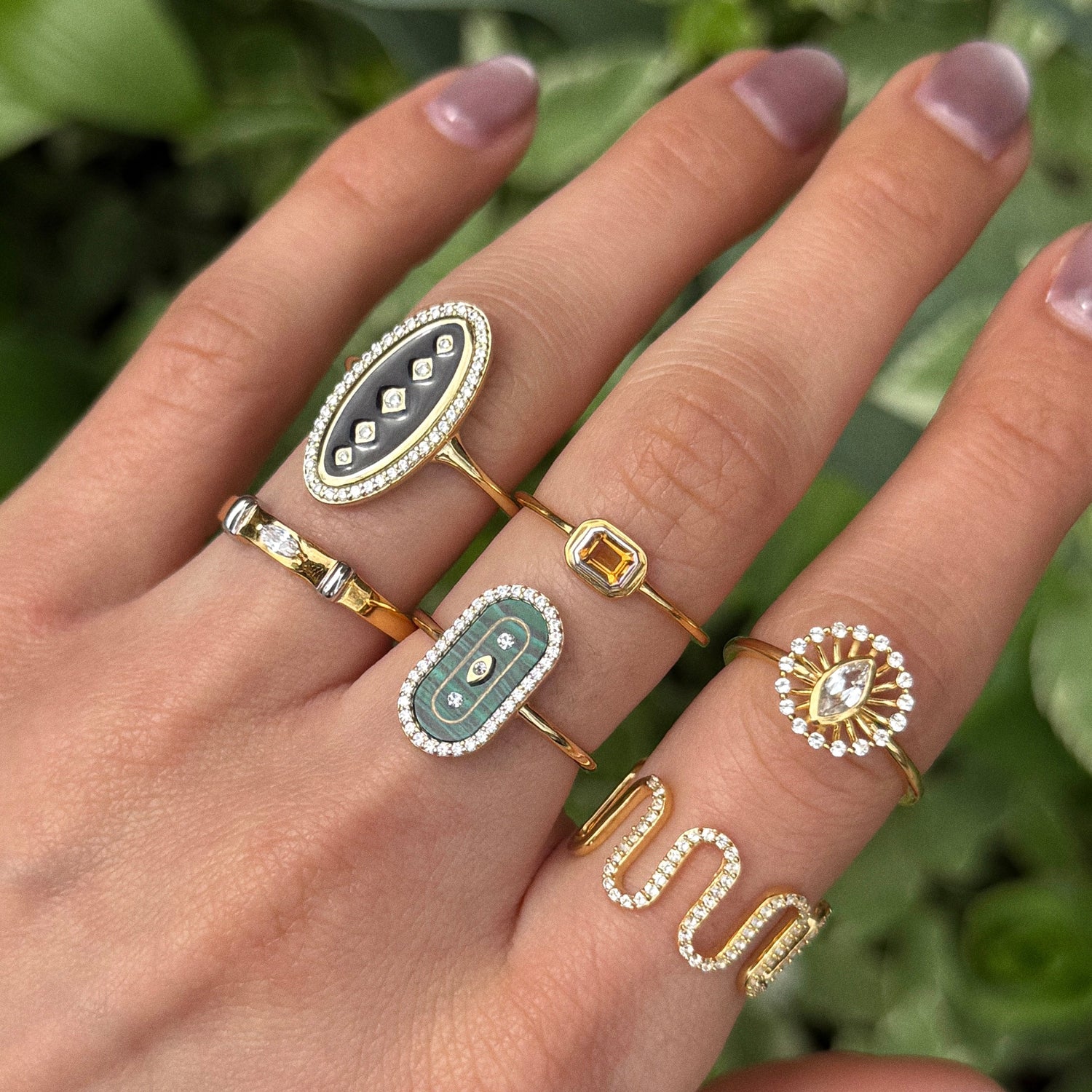 A hand with manicured nails displays the Velvet Noir Navette Ring—crafted in 14k yellow gold vermeil with navette shapes, CZ, black enamel accents, and diamond details—against a blurred green leafy background. Image